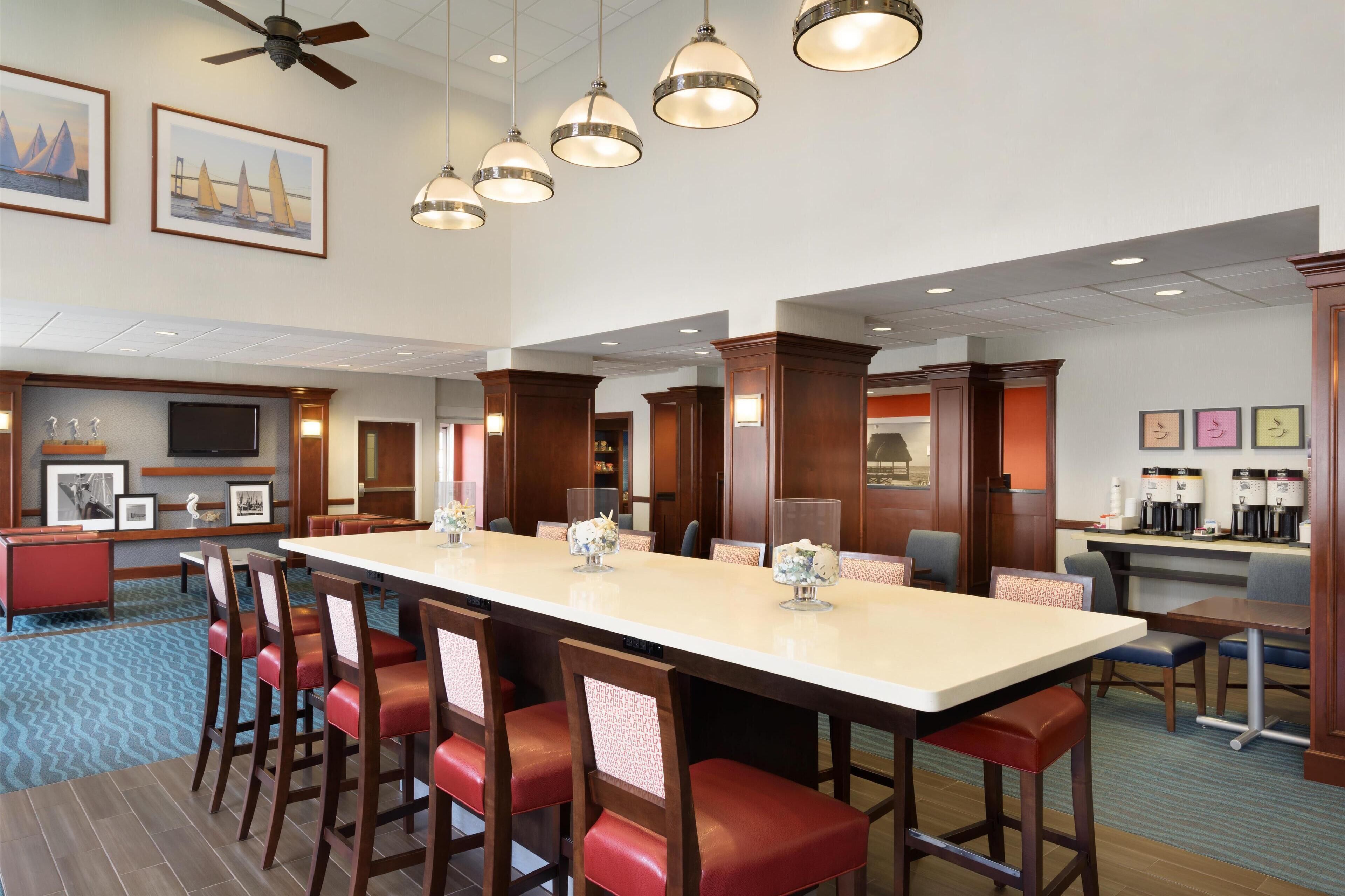 A hotel lobby with a long communal table, red cushioned chairs, a coffee station, and framed artwork on the walls.
