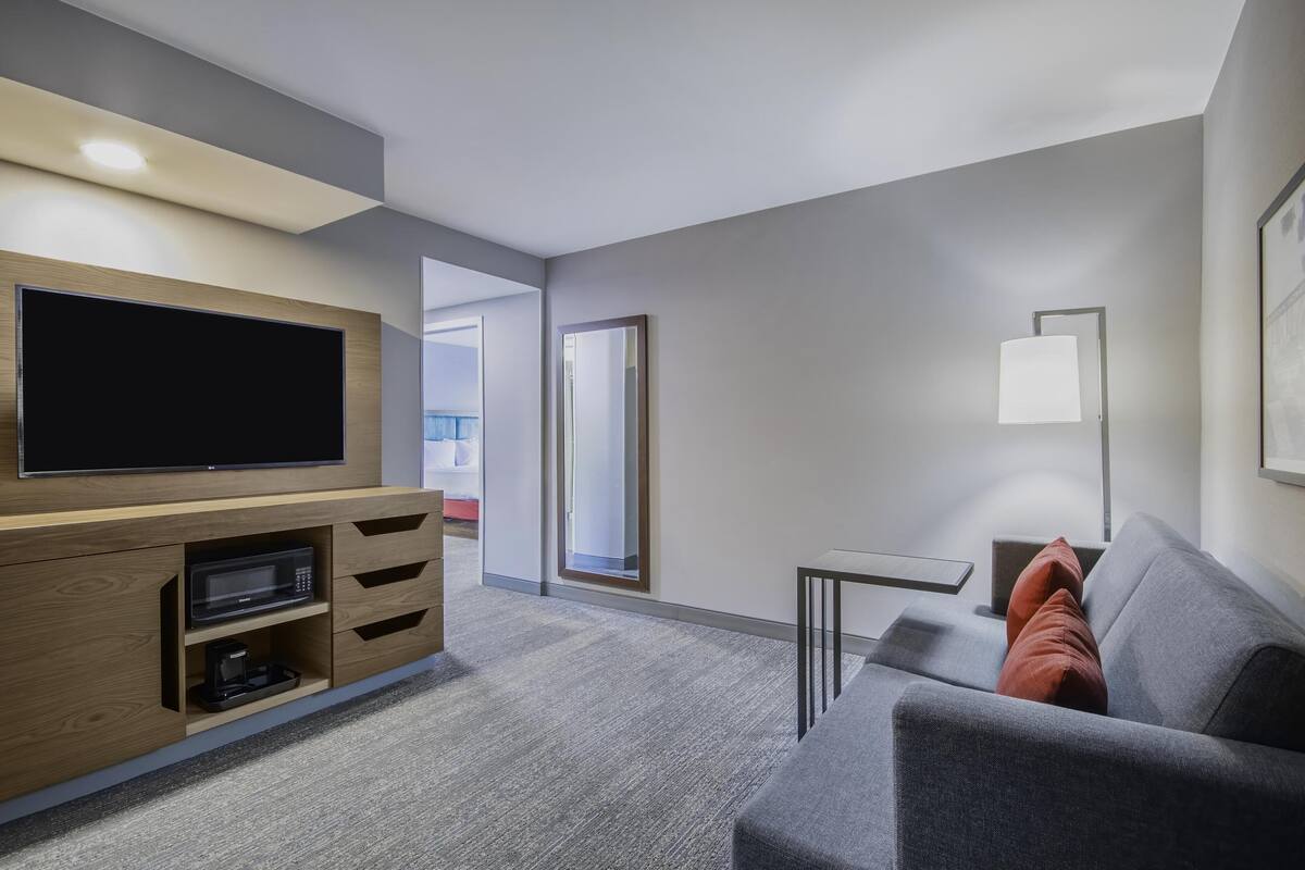 A modern hotel room with a flat-screen TV, a wooden entertainment unit, a grey sofa, and a small table with a lamp.