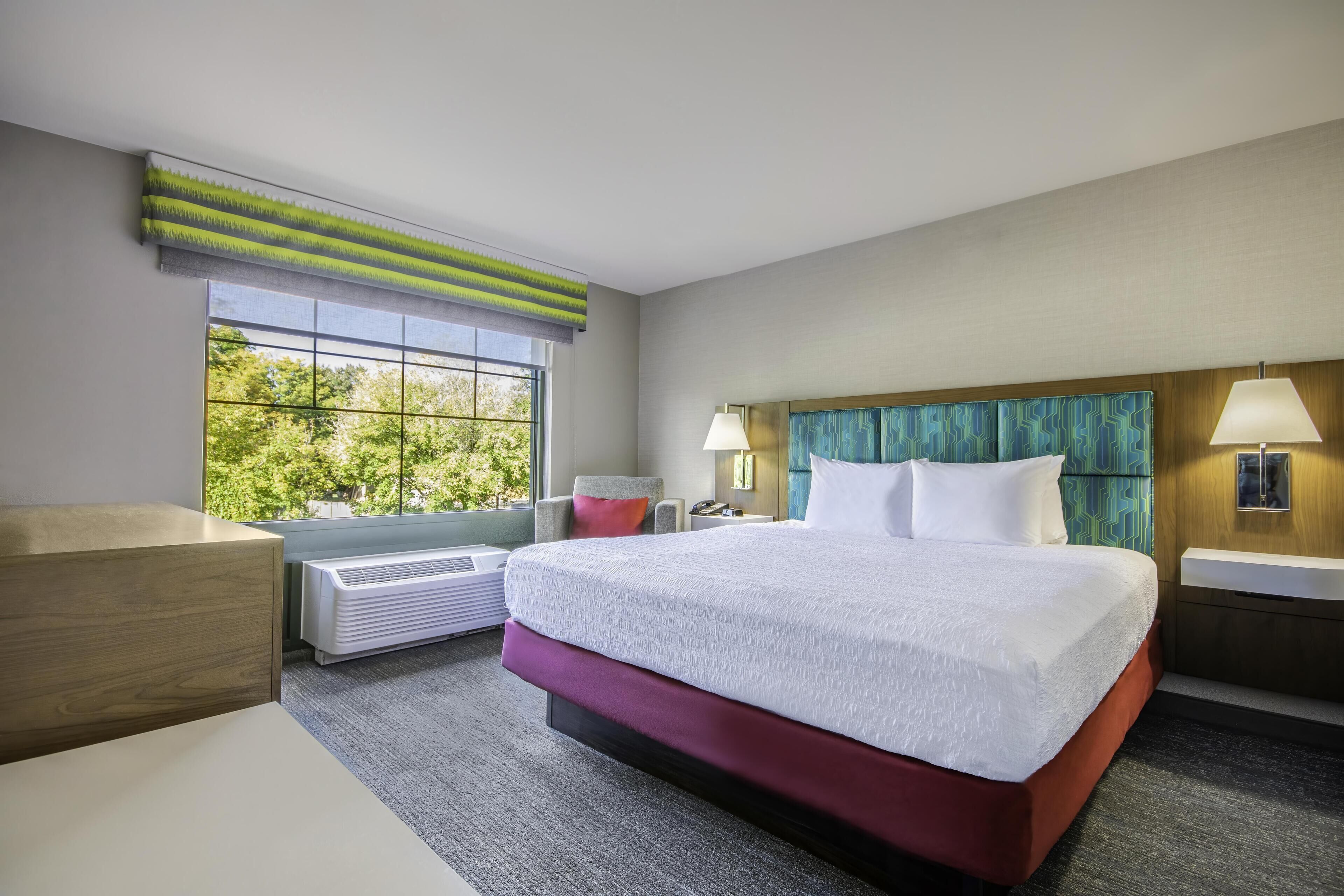 A hotel room with a large bed, a desk, a chair, a window with a view of trees, and a wall-mounted lamp.