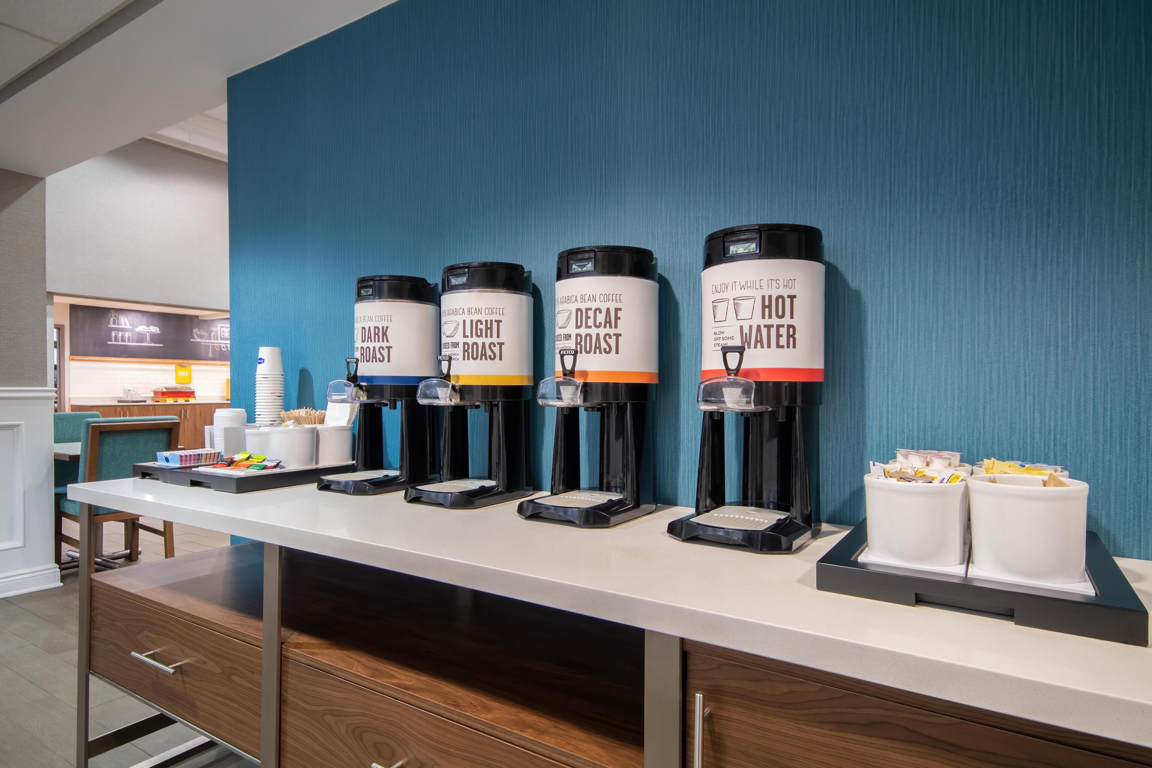 Un espace dédié au café, équipé de quatre distributeurs étiquetés « Café torréfié foncé », « Café torréfié léger », « Café décaféiné » et « Eau chaude ».