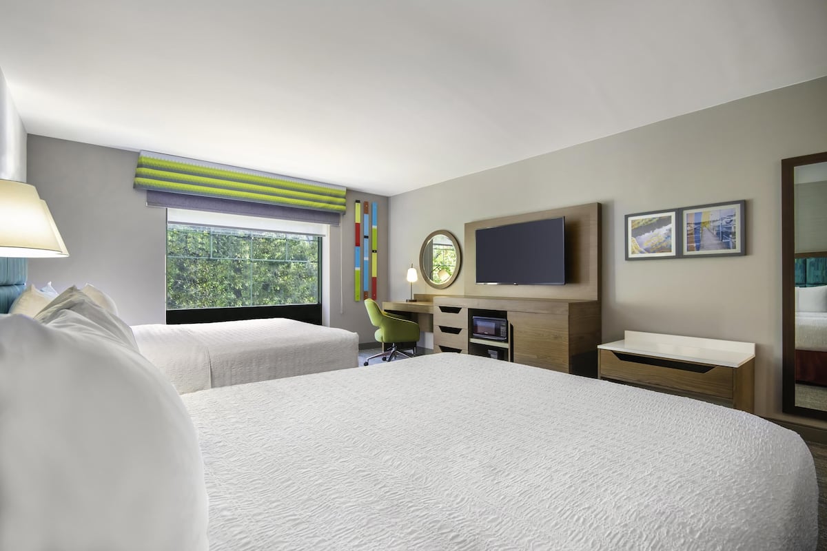 A hotel room with a large bed, two smaller beds, a desk with a computer, a TV, and a window with a view of greenery.