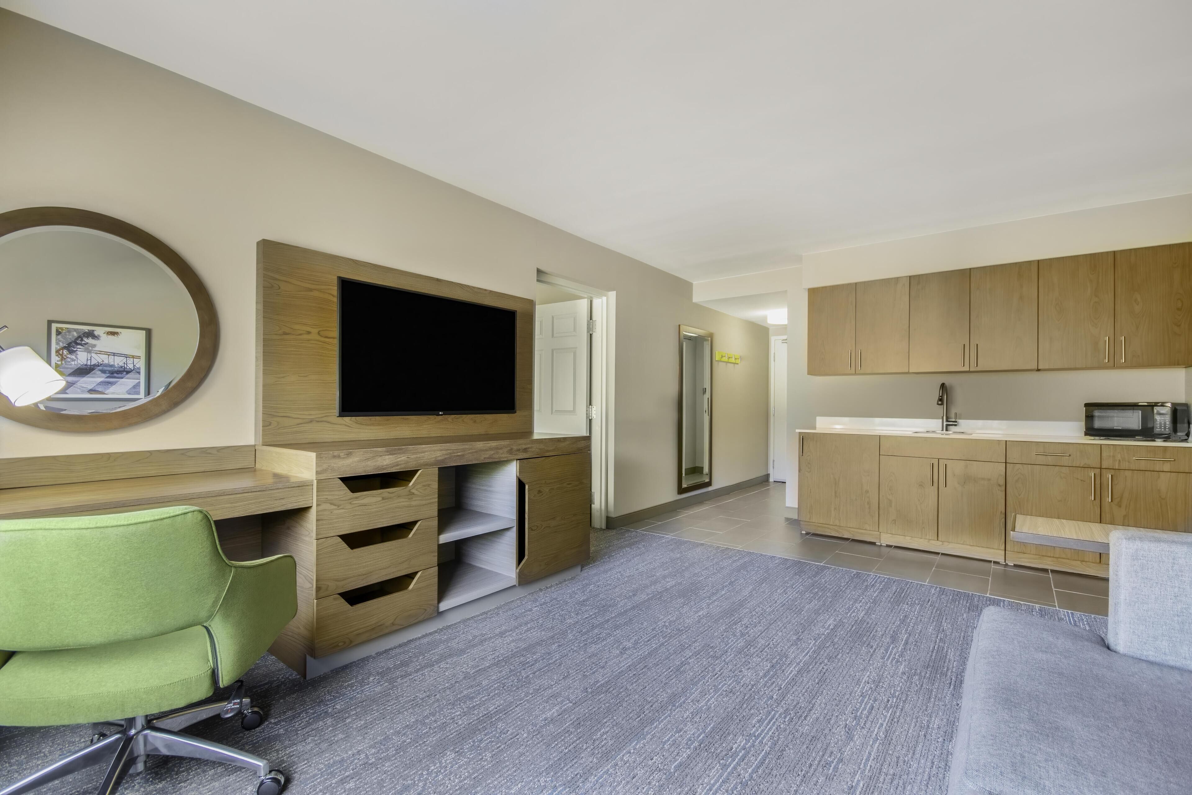 Een moderne hotelkamer met een flatscreen televisie, een groene bureaustoel, een grijze bank, houten kasten en een kitchenette.