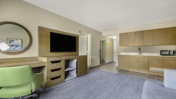 Een moderne hotelkamer met een flatscreen televisie, een groene bureaustoel, een grijze bank, houten kasten en een kitchenette.