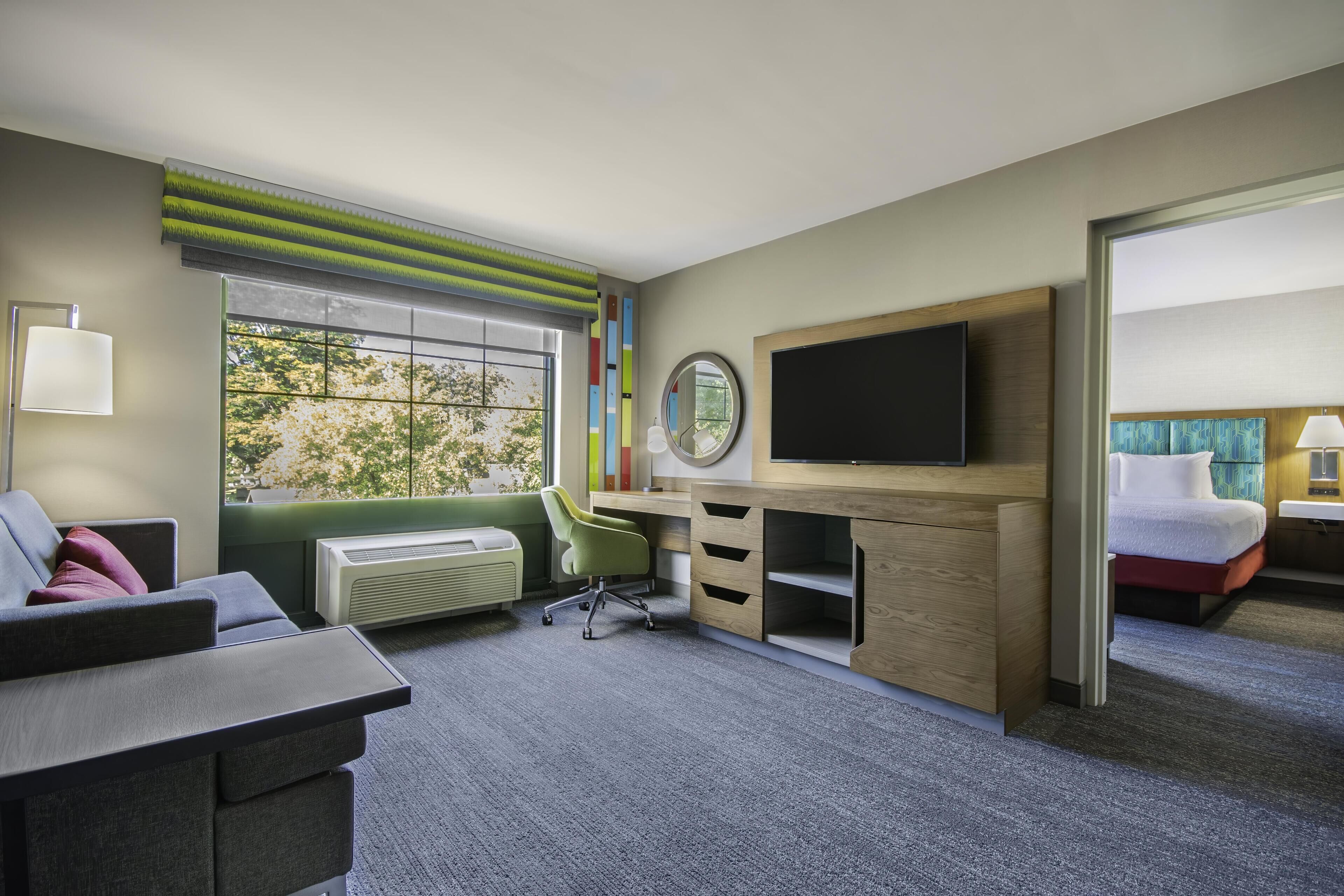 A hotel room with a desk, chair, TV, bed, and a window with a view of trees.