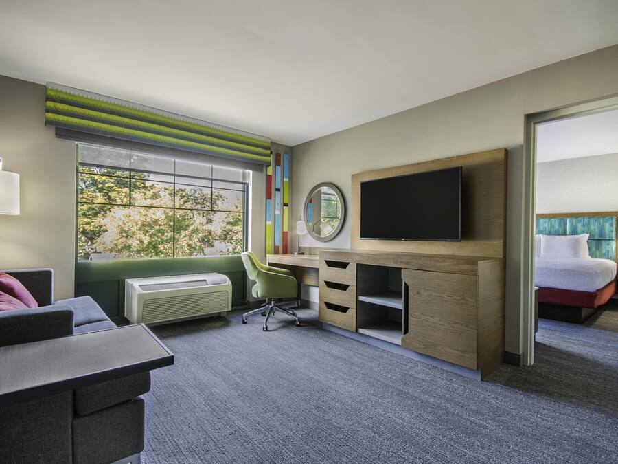 A hotel room with a desk, chair, TV, bed, and a window with a view of trees.