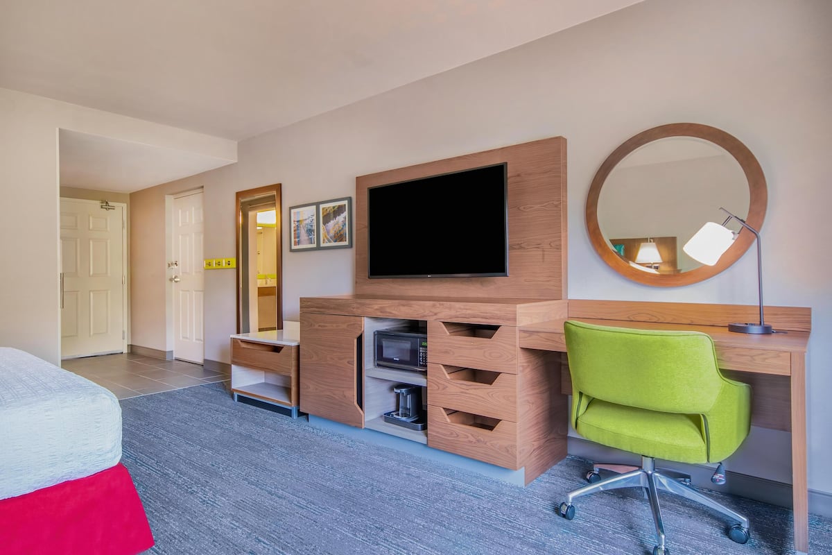 A hotel room with a bed, a desk with a flat-screen TV, a mirror, a green office chair, and a coffee maker.