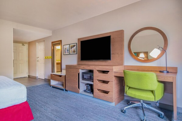 A hotel room with a bed, a desk with a flat-screen TV, a mirror, a green office chair, and a coffee maker.