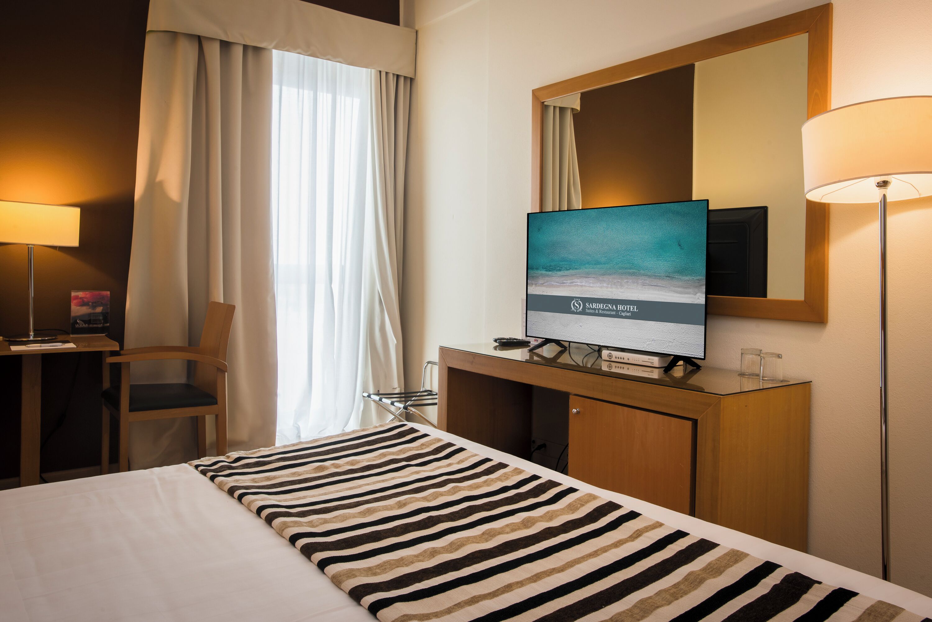 Una habitación de hotel con cama, escritorio, silla, televisor y espejo.