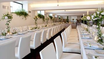 Une longue table à manger, entourée de chaises blanches, dressée avec de la vaisselle et des verres, et agrémentée de compositions florales et de bougies.