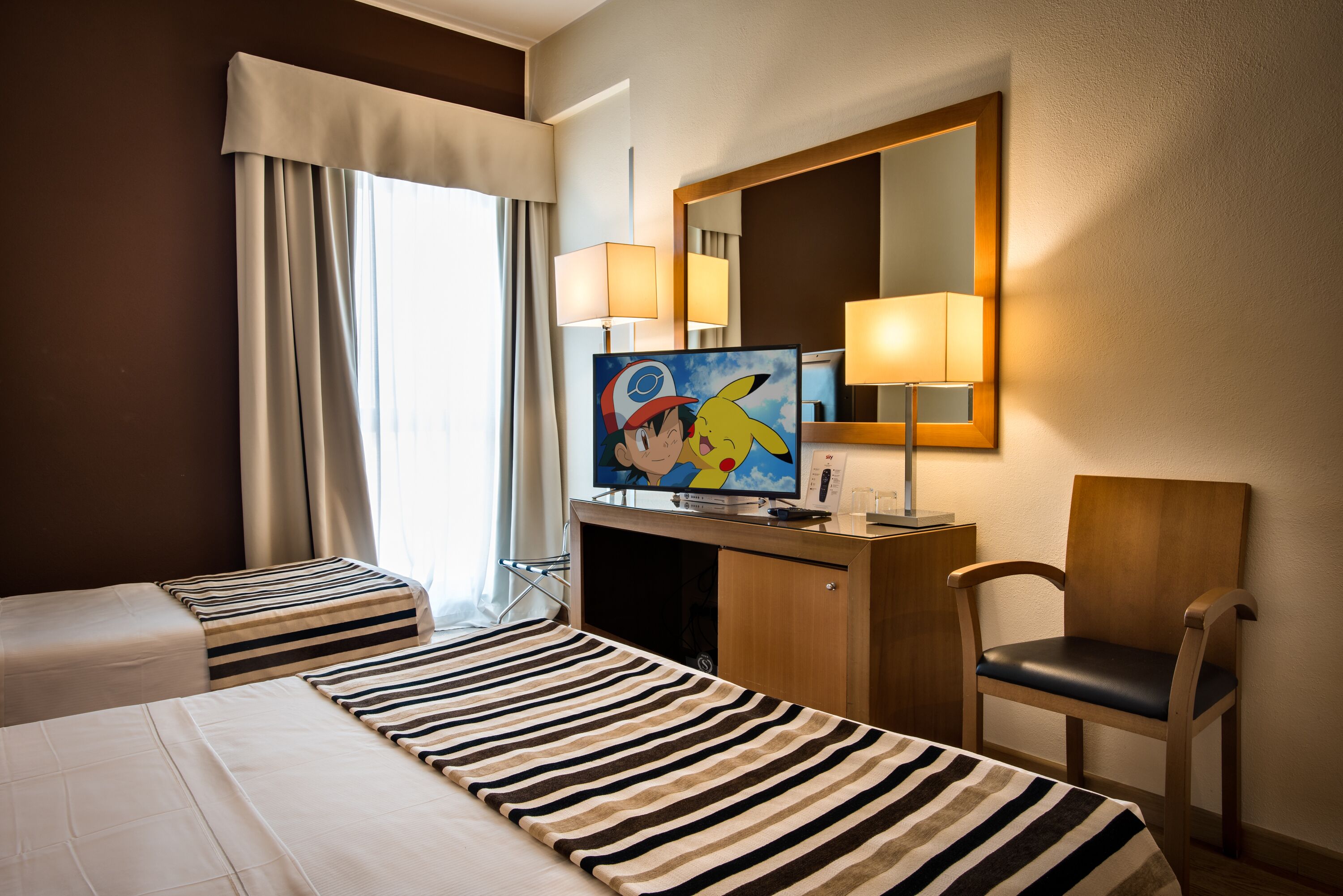 A hotel room with two beds, a television, a desk, a chair, and a large mirror.
