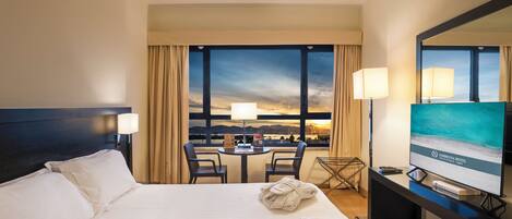 A hotel room with a bed, a desk with a TV, a chair, and a view of the sunset through large windows.