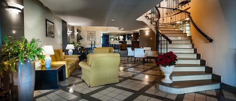 A hotel lobby with a checkered floor, a staircase with a metal railing, and seating areas with lamps and plants.