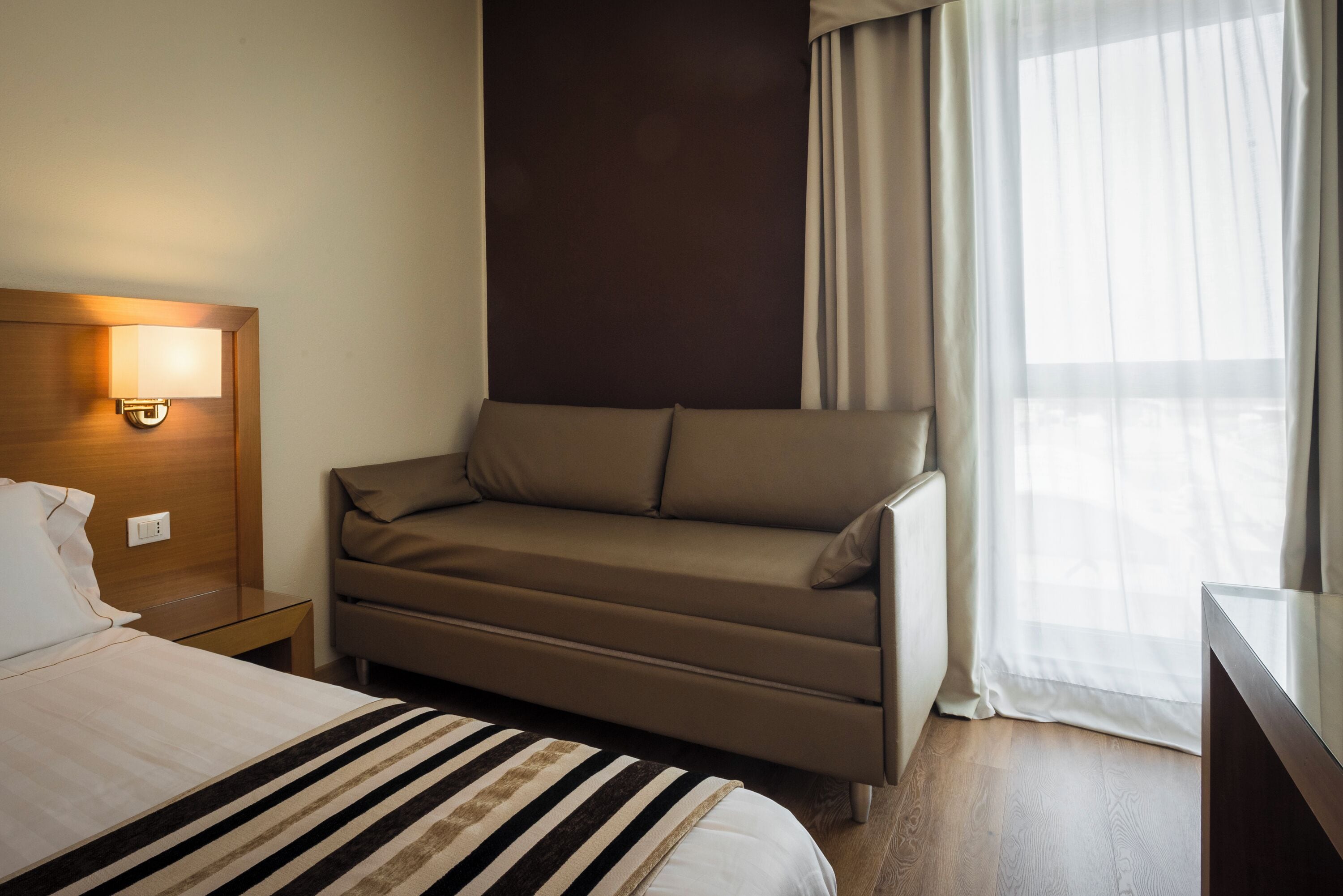 Una habitación de hotel con cama, sofá, mesita de noche con lámpara y ventana con cortinas.