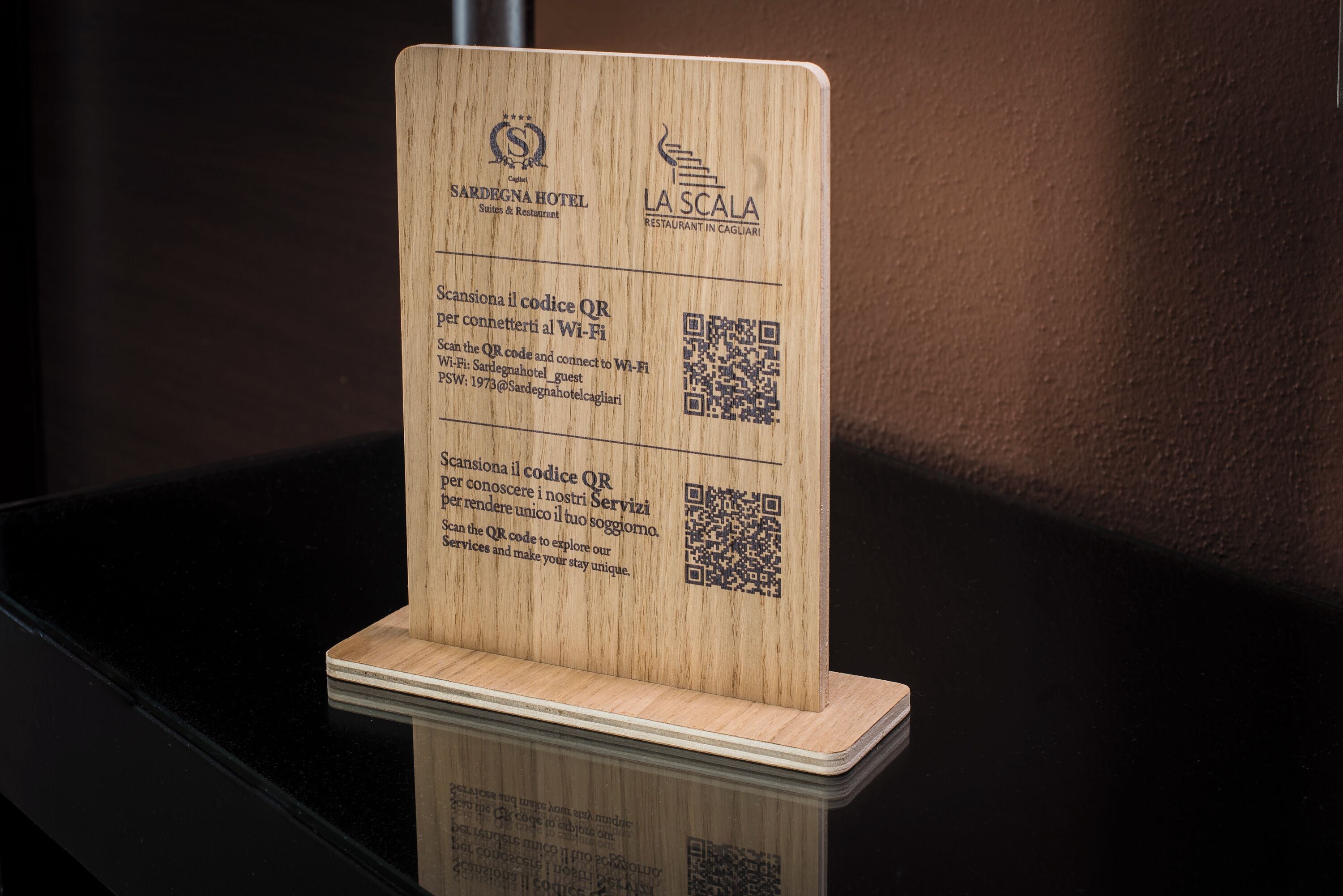 A wooden sign with QR codes and text in Italian, promoting Sardegna Hotel and La Scala Restaurant in Cagliari.