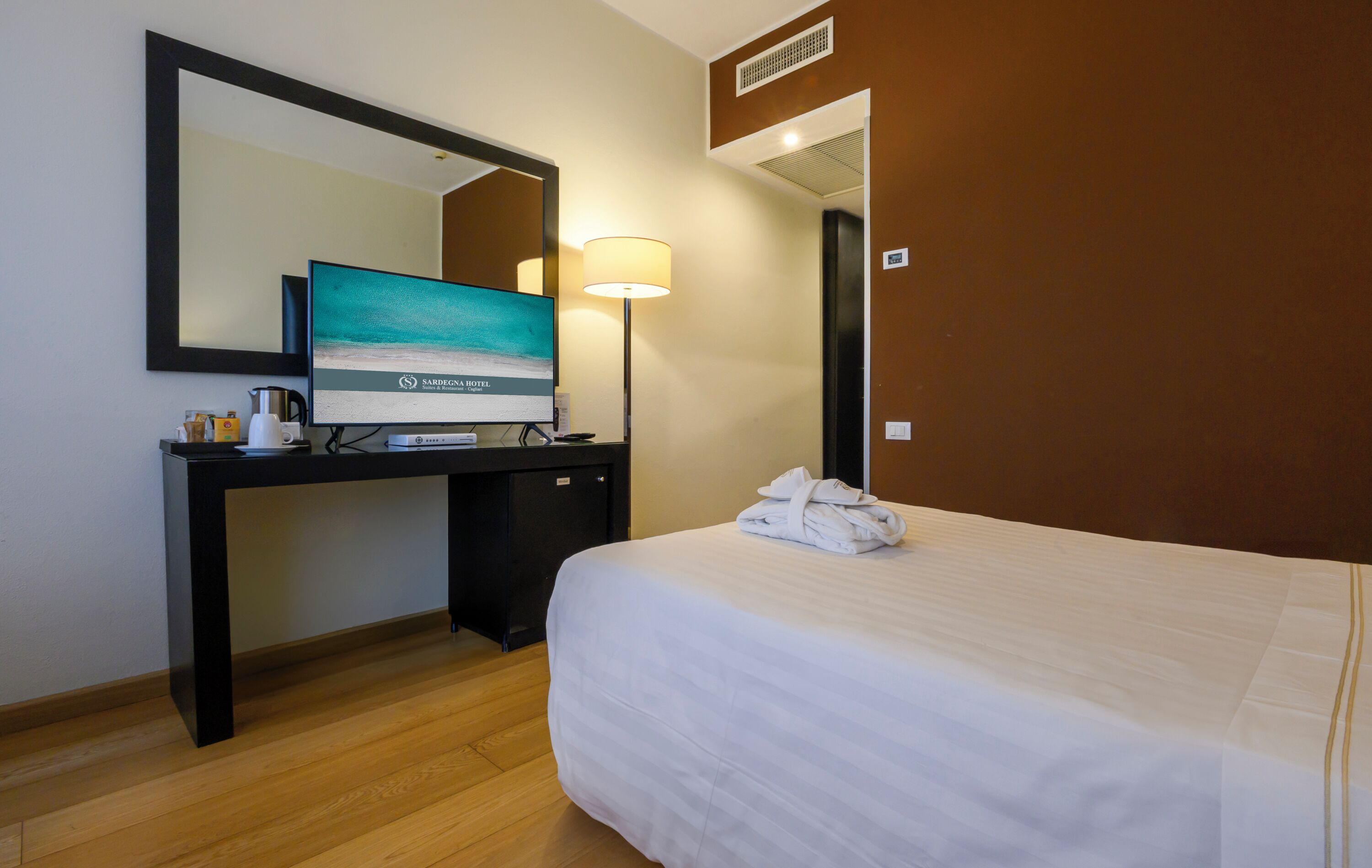 A hotel room with a bed, a desk with a computer monitor, a lamp, and a mirror.