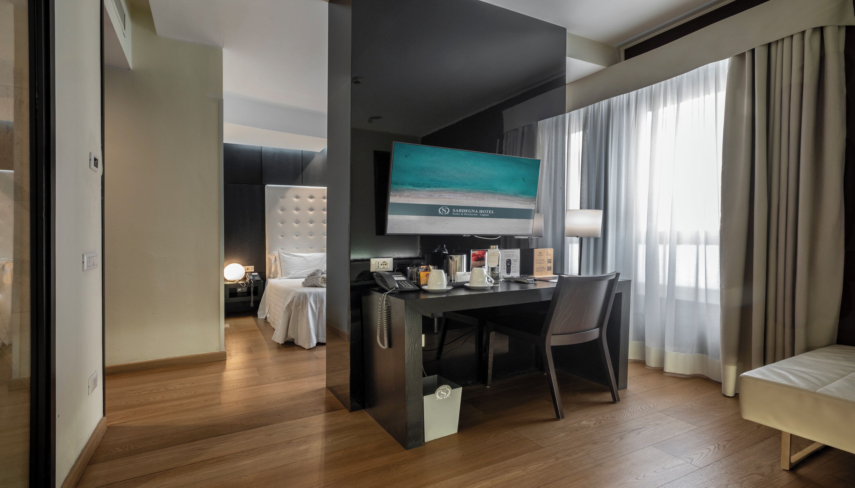 Una habitación de hotel con cama, escritorio, computadora, televisor y ventana con cortinas.