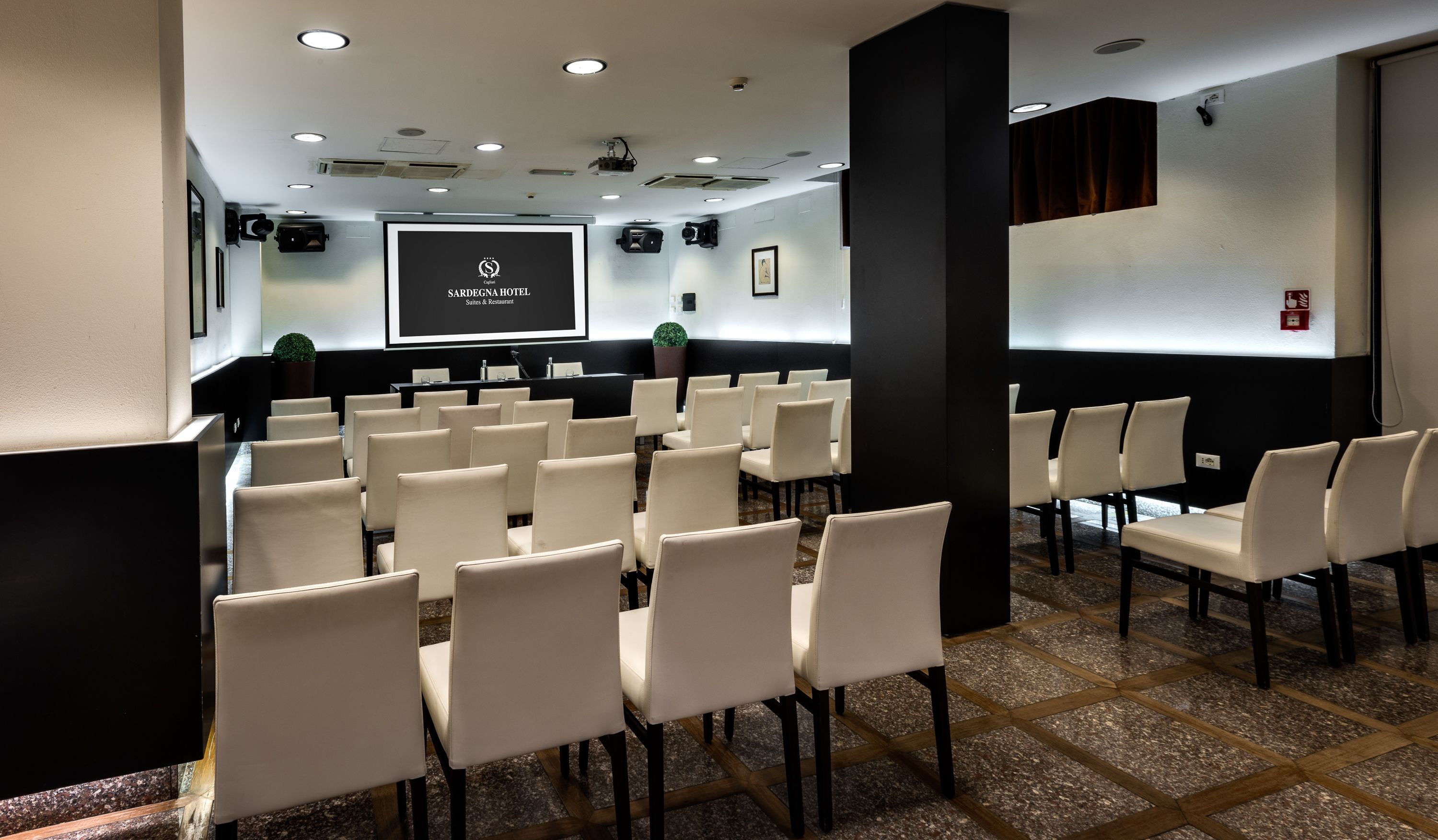Un salón de conferencias con filas de sillas blancas, una pantalla de proyector y un atril negro.