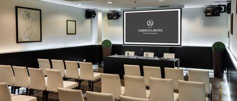 A conference room with a projector screen displaying the Sardegna Hotel logo, a black table with chairs, and framed artwork on the walls.