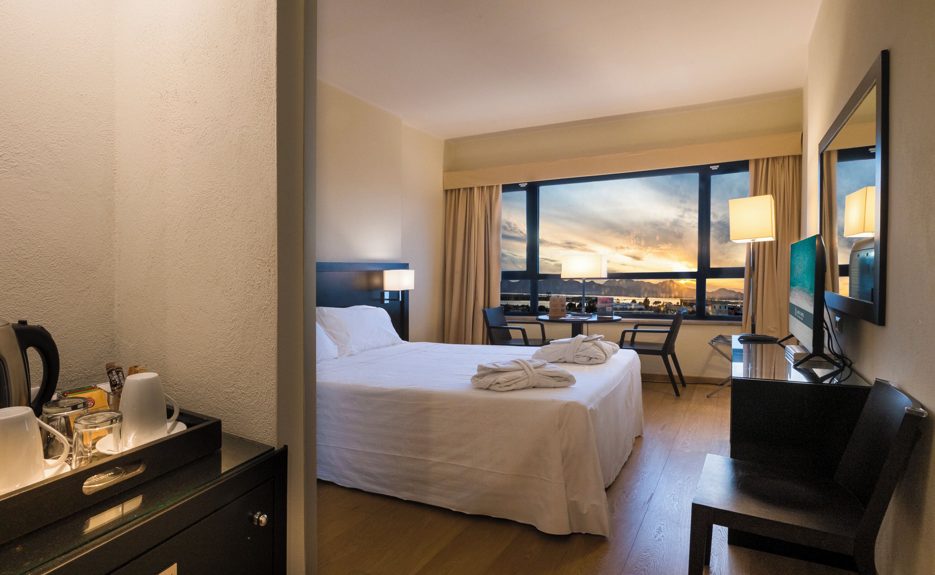 A hotel room with a bed, two chairs, a desk, a TV, and a window with a sunset view.