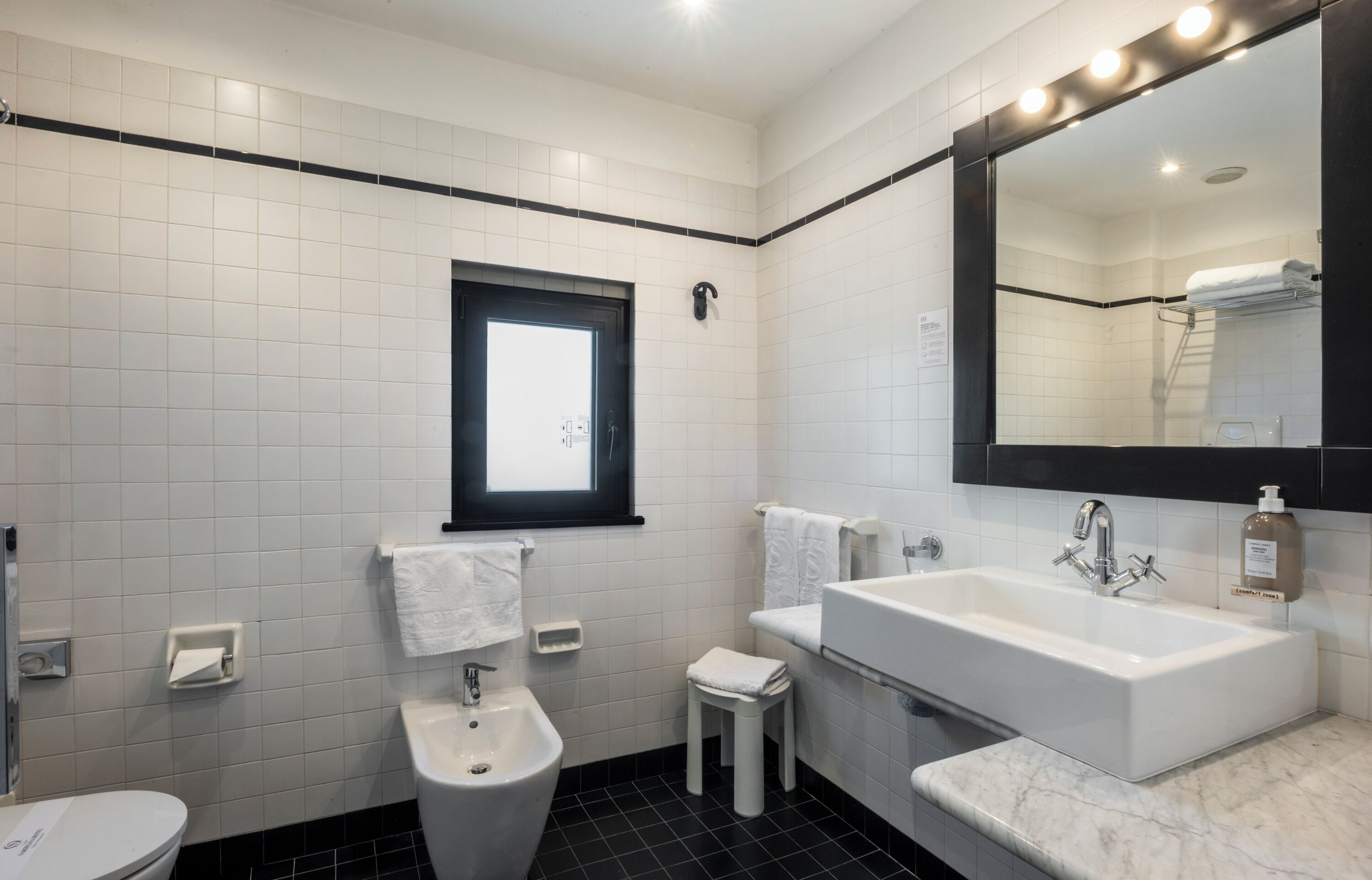 A modern bathroom with a large mirror, white tiles, a black floor, a toilet, a bidet, and a sink.