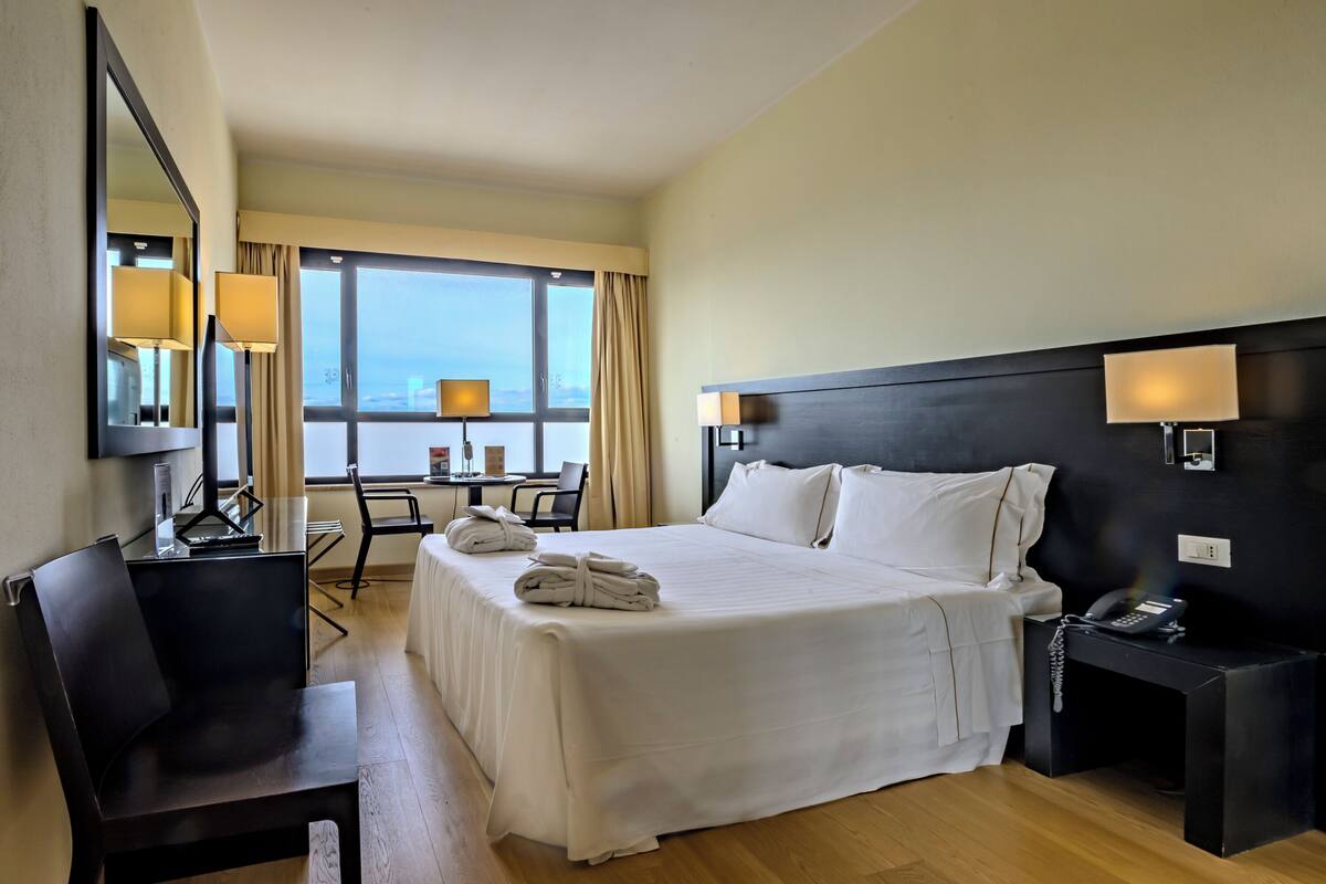 Une chambre d’hôtel avec un grand lit, un bureau avec une chaise, une petite table et une vue sur la ville par la fenêtre.