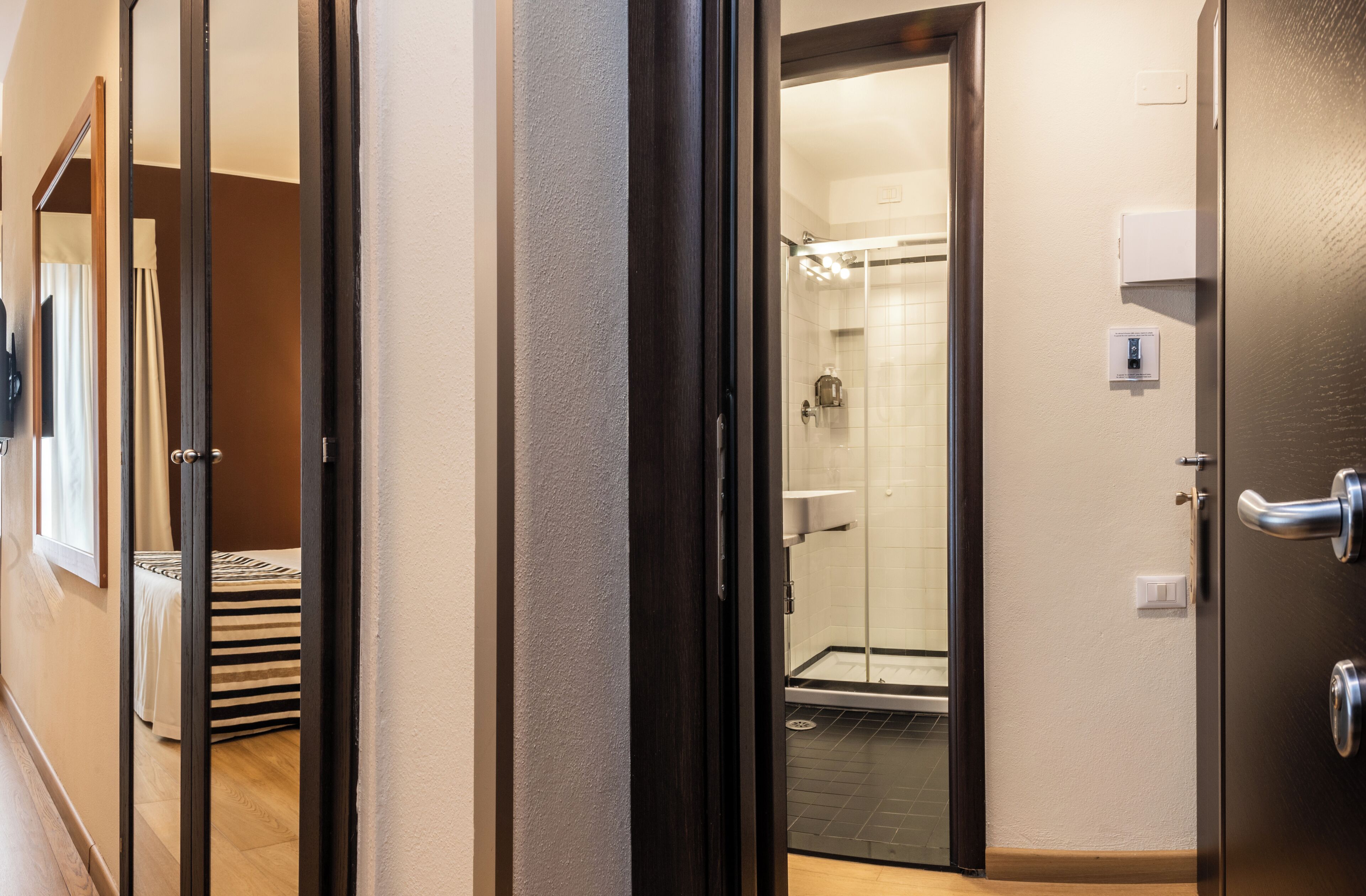 A hotel room with a glass shower door, a bed with striped bedding, and a wooden floor.