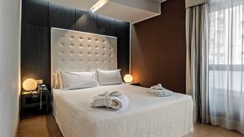 Habitación de hotel moderna con una cama grande, ropa de cama blanca, cabecero acolchado, lámparas de noche y vistas al exterior a través de cortinas de gasa.
