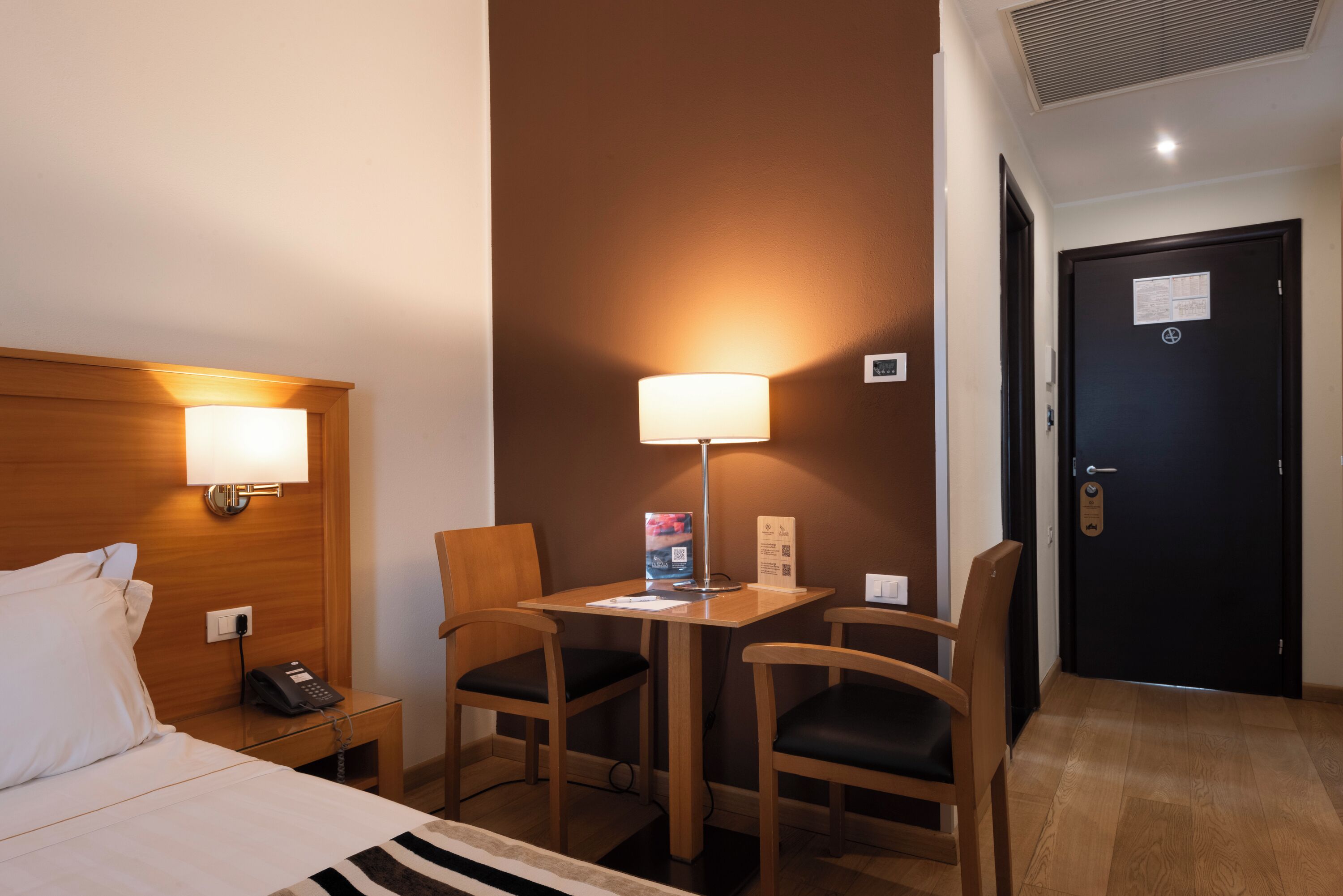 Habitació d'hotel amb llit, làmpada de nit, escriptori amb làmpada, dues cadires i una porta.