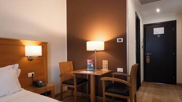 A hotel room with a bed, bedside lamp, desk with a lamp, two chairs, and a door.