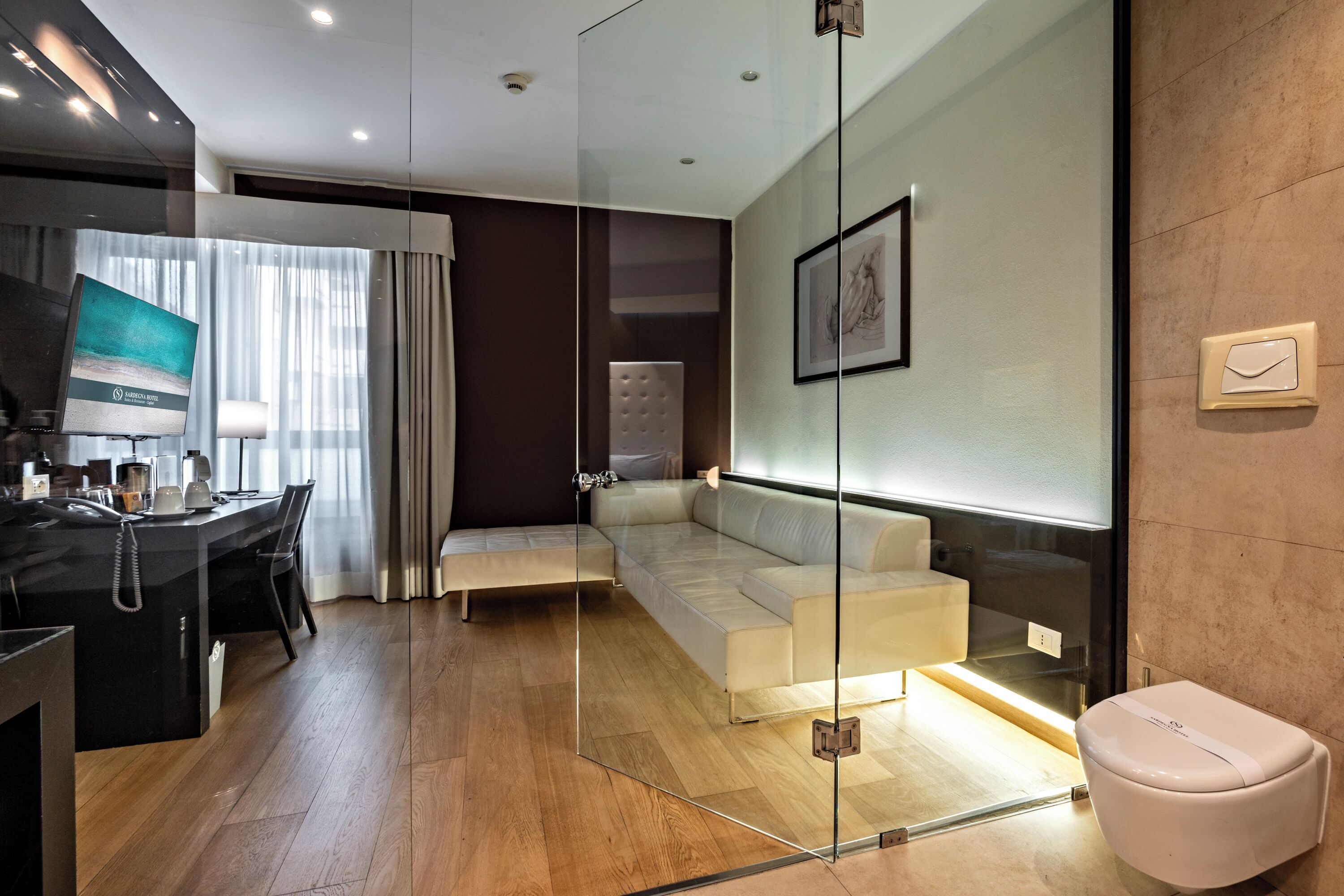 Quarto de hotel moderno com divisória de vidro, sofá, escrivaninha com telefone e banheiro com vaso sanitário.