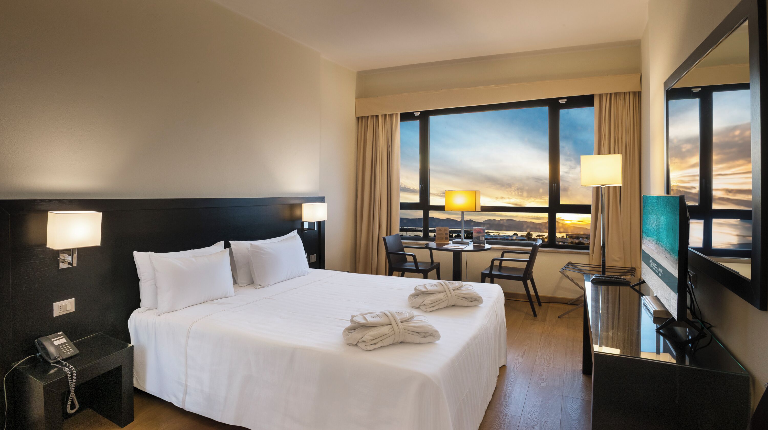 Ein Hotelzimmer mit einem großen Bett, Nachttischlampen, einem Schreibtisch mit Stuhl, einem Fernseher und Blick auf den Sonnenuntergang durch das Fenster.