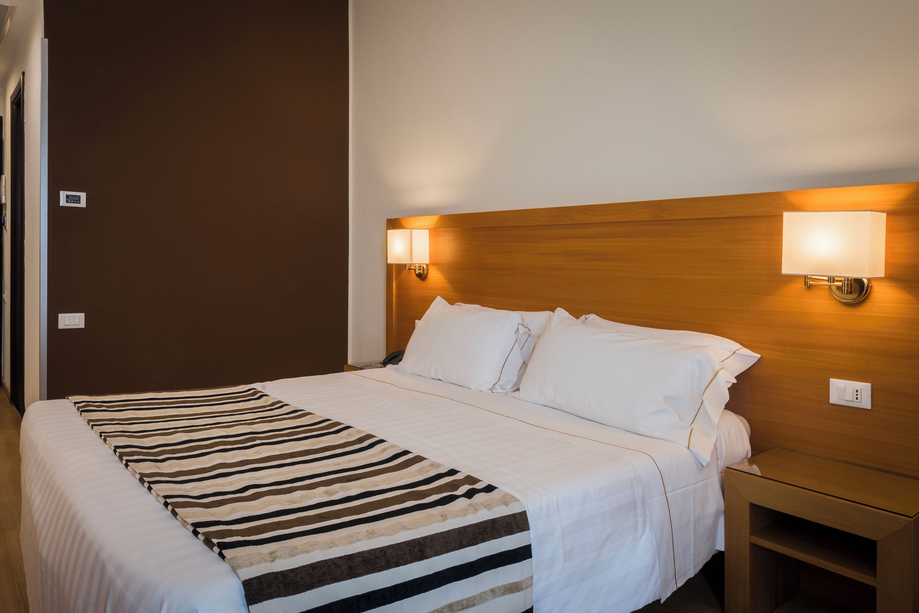 A hotel room with a large bed, a wooden headboard, two bedside lamps, and a bedside table.