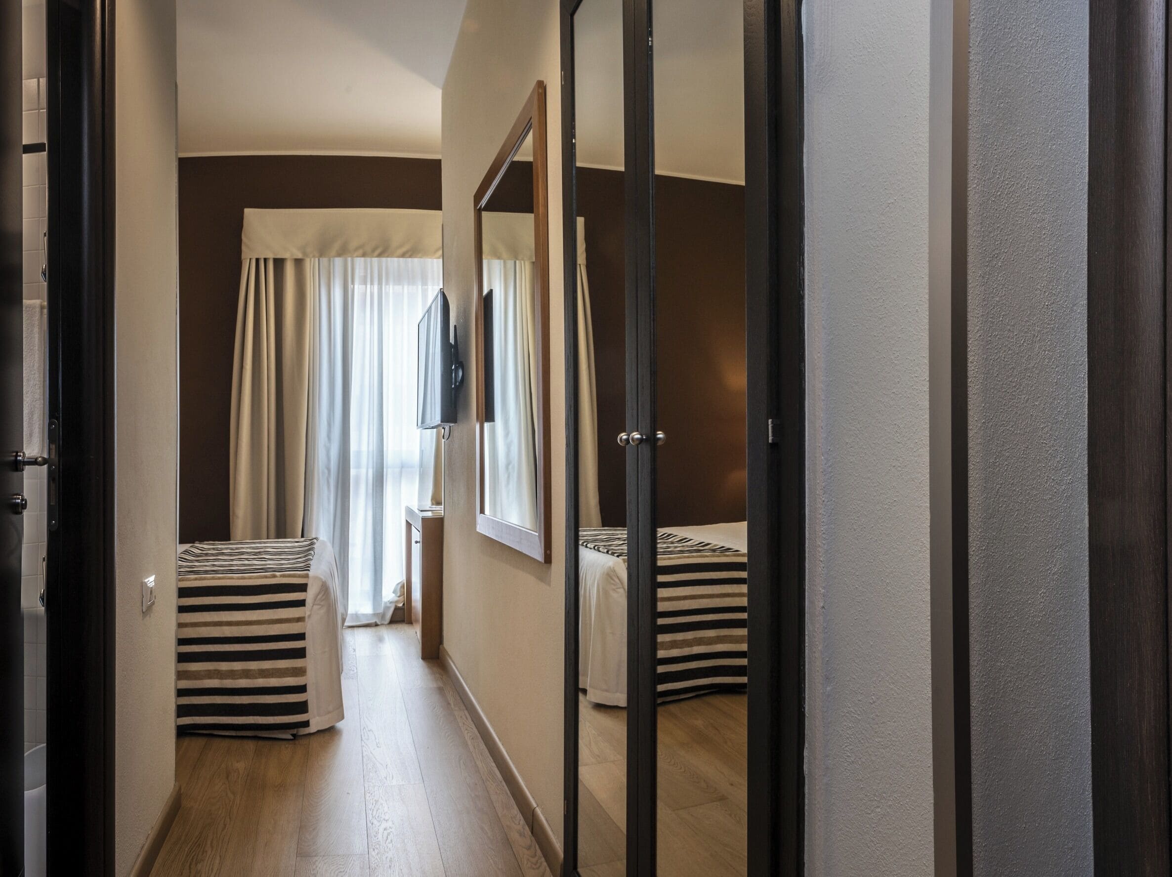 A hotel room with a wooden floor, a bed with striped bedding, a large mirror, and a window with curtains.