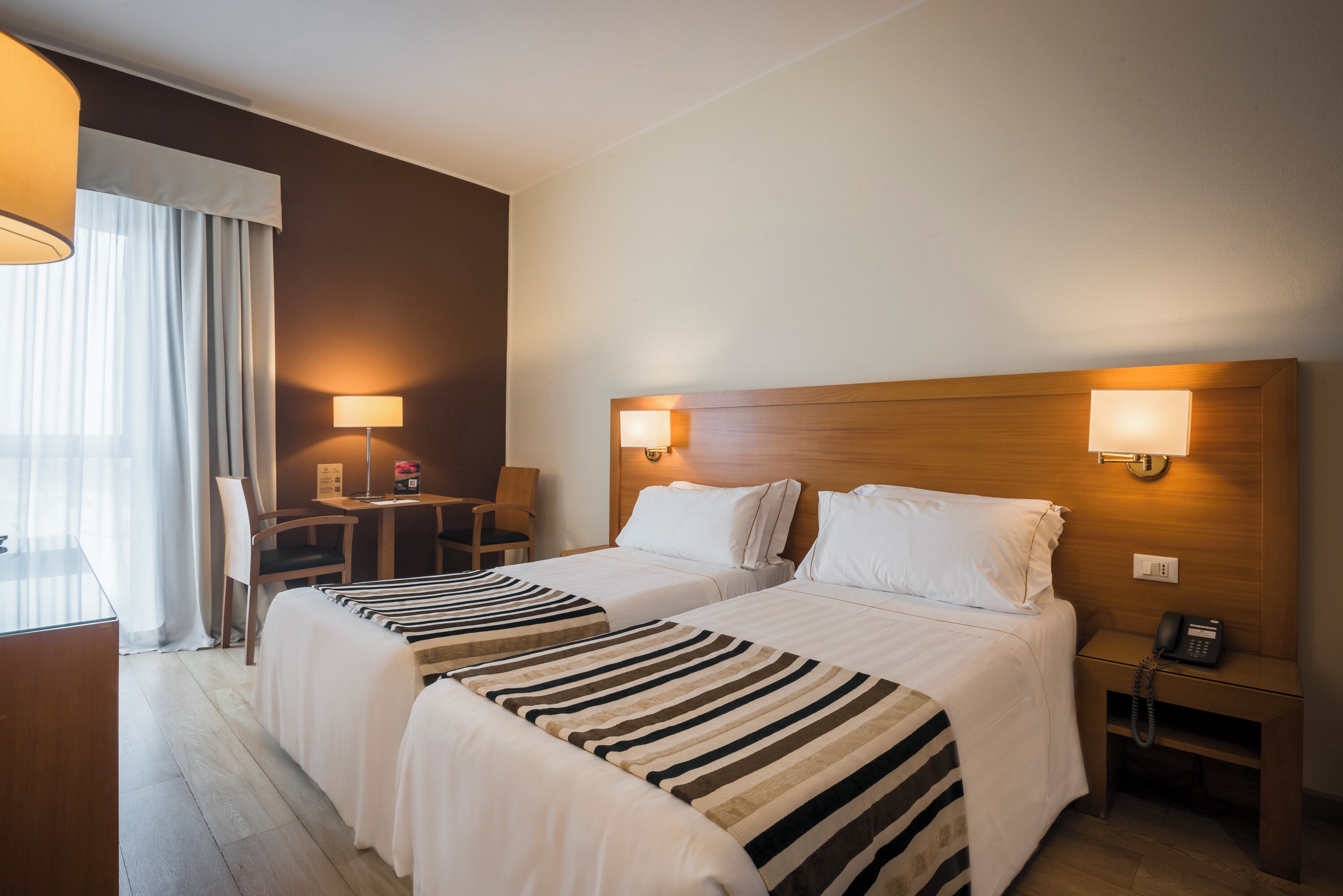 Una camera d'albergo con due letti, una scrivania, una sedia, una lampada e un telefono sul comodino.