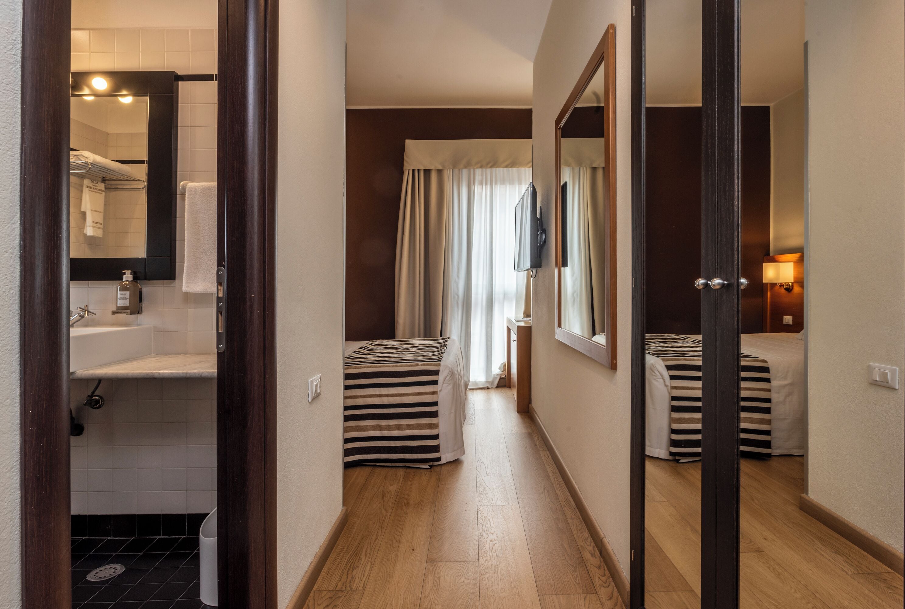 A hotel room with a wooden floor, a bed with striped bedding, a bedside table with a lamp, and a bathroom with a sink and mirror.