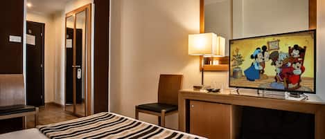 A hotel room with a bed, a desk with a lamp, a television displaying animated characters, and a chair.