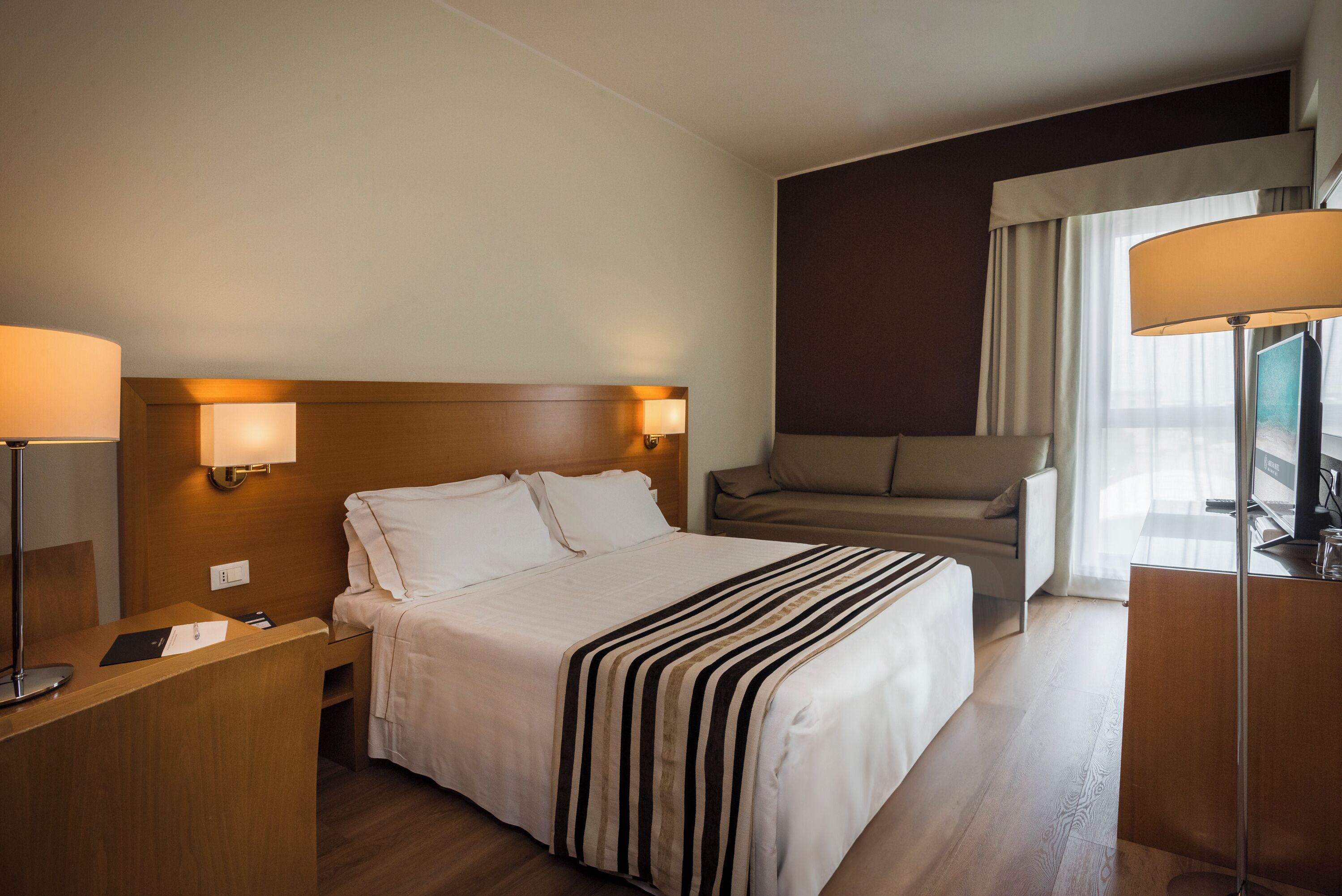 A hotel room with a bed, bedside tables, a desk, a chair, a sofa, and a TV.