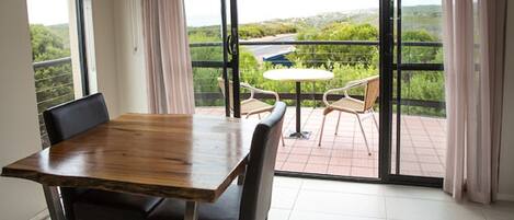 Un comedor con mesa y sillas de madera, ventanas amplias con vistas a la vegetación y una carretera, y un balcón con mesa y sillas.