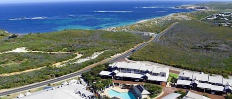 An aerial view of a coastal resort with a road leading to a cluster of white-roofed buildings, a swimming pool, and a view of the ocean.