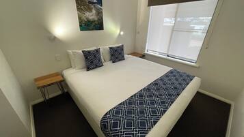 Una cama bien hecha con colcha blanca y azul marino, dos almohadas a juego en azul marino, una mesita de noche de madera, una lámpara de pared y una ventana con persianas.