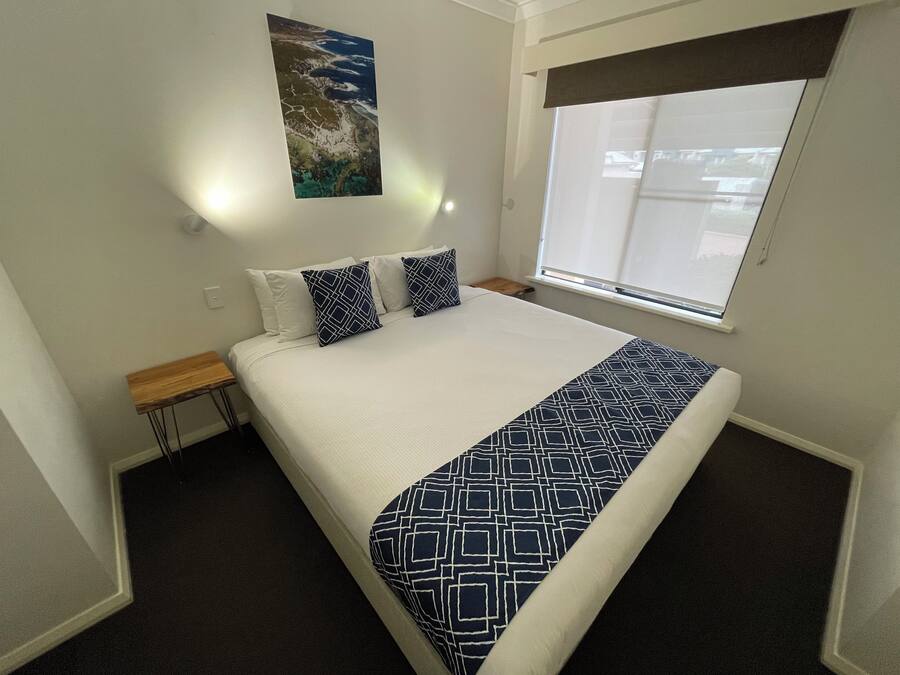 Una cama bien hecha con colcha blanca y azul marino, dos almohadas a juego en azul marino, una mesita de noche de madera, una lámpara de pared y una ventana con persianas.