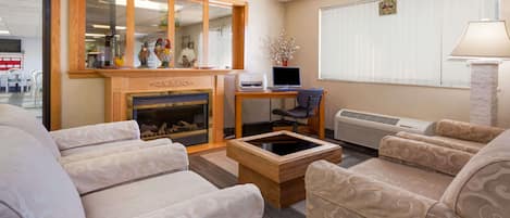 Una sala de estar con sofás color beige con estampado, una mesa de centro de madera, una chimenea, un escritorio con ordenador y un ventanal con persianas.