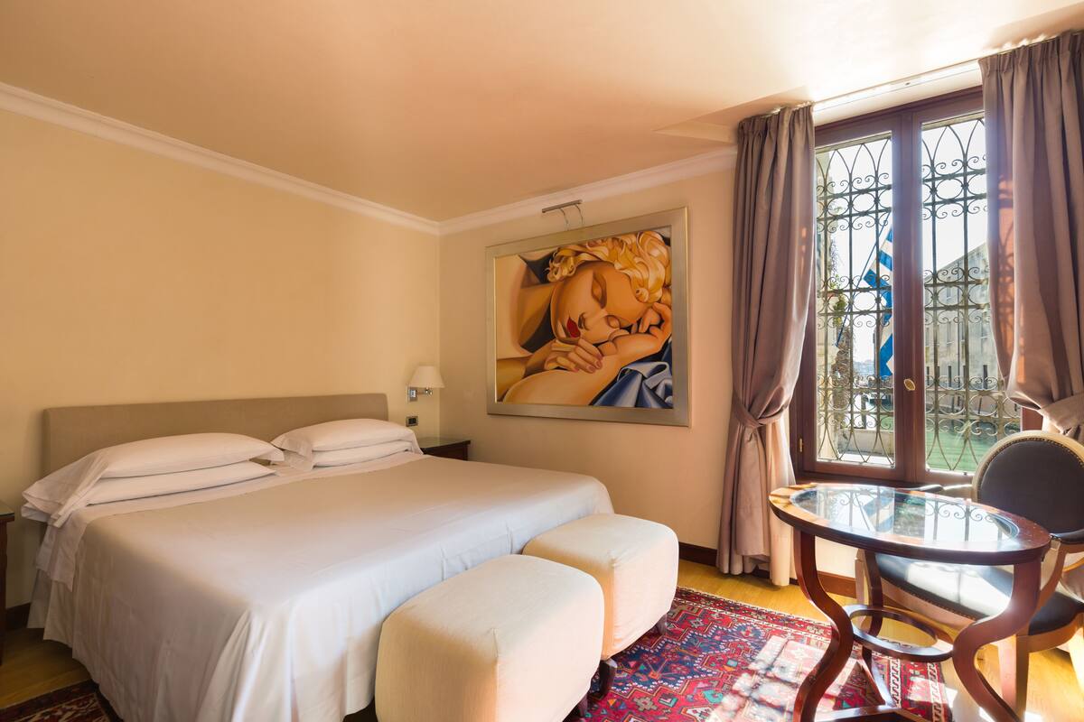 Ein Hotelzimmer mit einem Bett, zwei Nachttischen, einem Sessel, einem Gemälde und einem fenster mit Vorhängen.