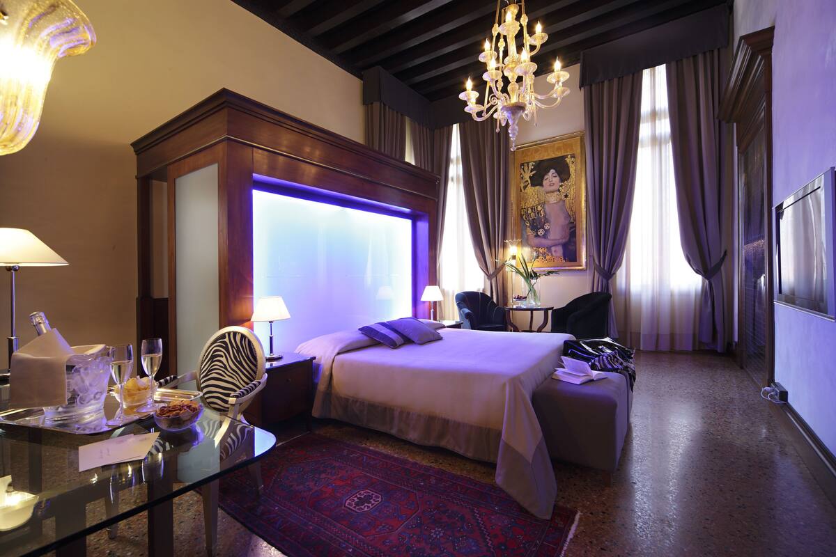 Ein Hotelzimmer mit einem großen Bett, einer Sitzecke, einem Schreibtisch mit Lampe, einem Kronleuchter und einem großen Fenster mit Vorhängen.