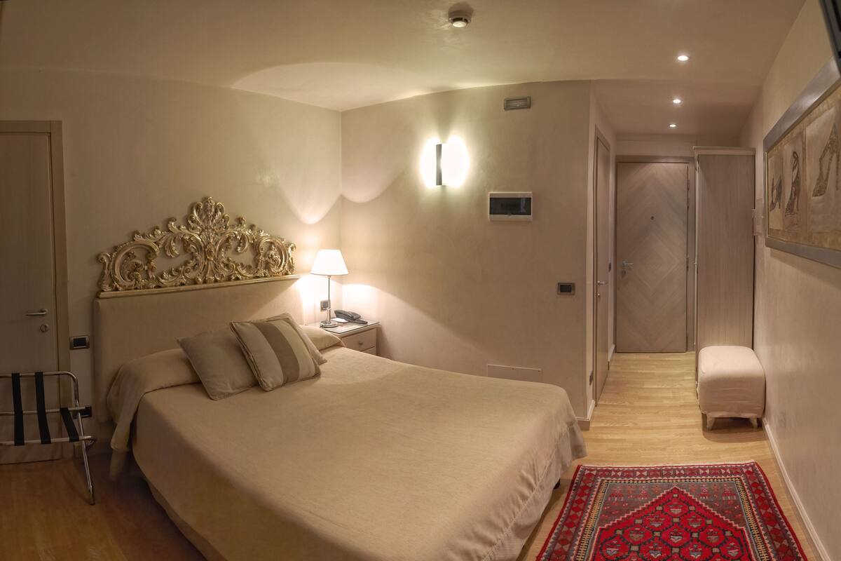 Ein Hotelzimmer mit einem großen Bett, einer Nachttischlampe und einem dekorativen Kopfteil. Ein kleiner Tisch mit Telefon und einer Wandleuchte steht ebenfalls dort.