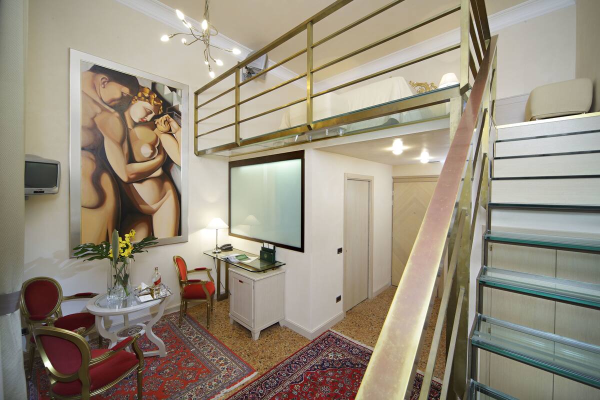 Ein Hotelzimmer mit hoher Decke, einer Glasstiege, einem großen Gemälde an der Wand, einem Schreibtisch mit Computer und einem roten Teppich.