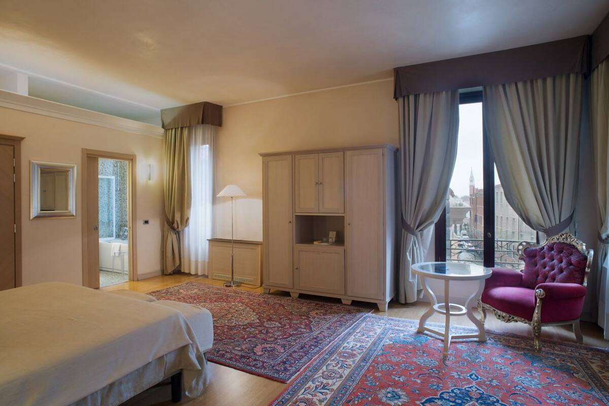 Ein Hotelzimmer mit Bett, Sitzecke mit Stuhl, kleinem Tisch, Kleiderschrank und Blick auf die Stadt durch das Fenster.