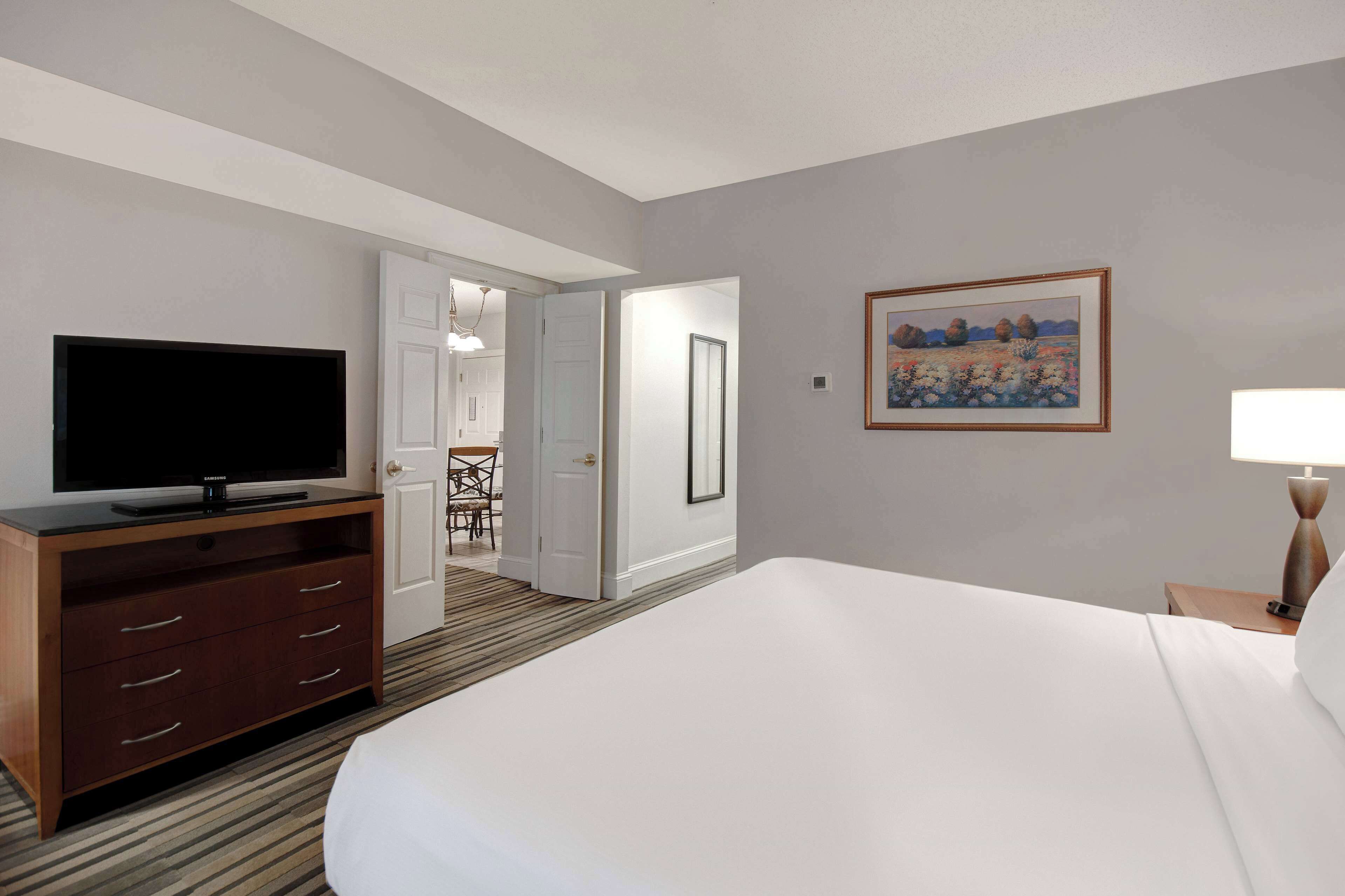 A hotel room with a large bed, a TV on a wooden dresser, a painting on the wall, and a door leading to a dining area.