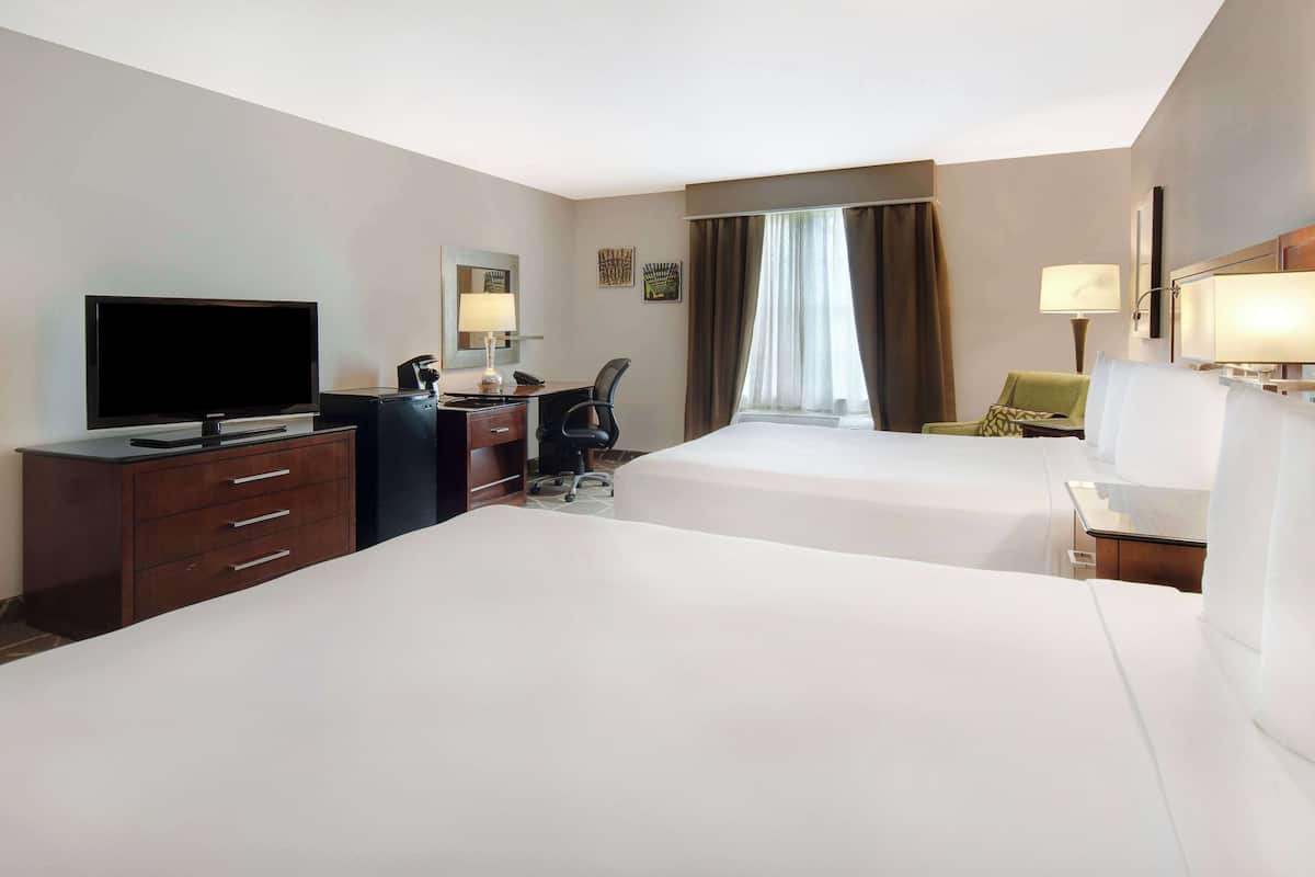 A hotel room with two beds, a desk with a flat-screen TV, a chair, and a window with curtains.