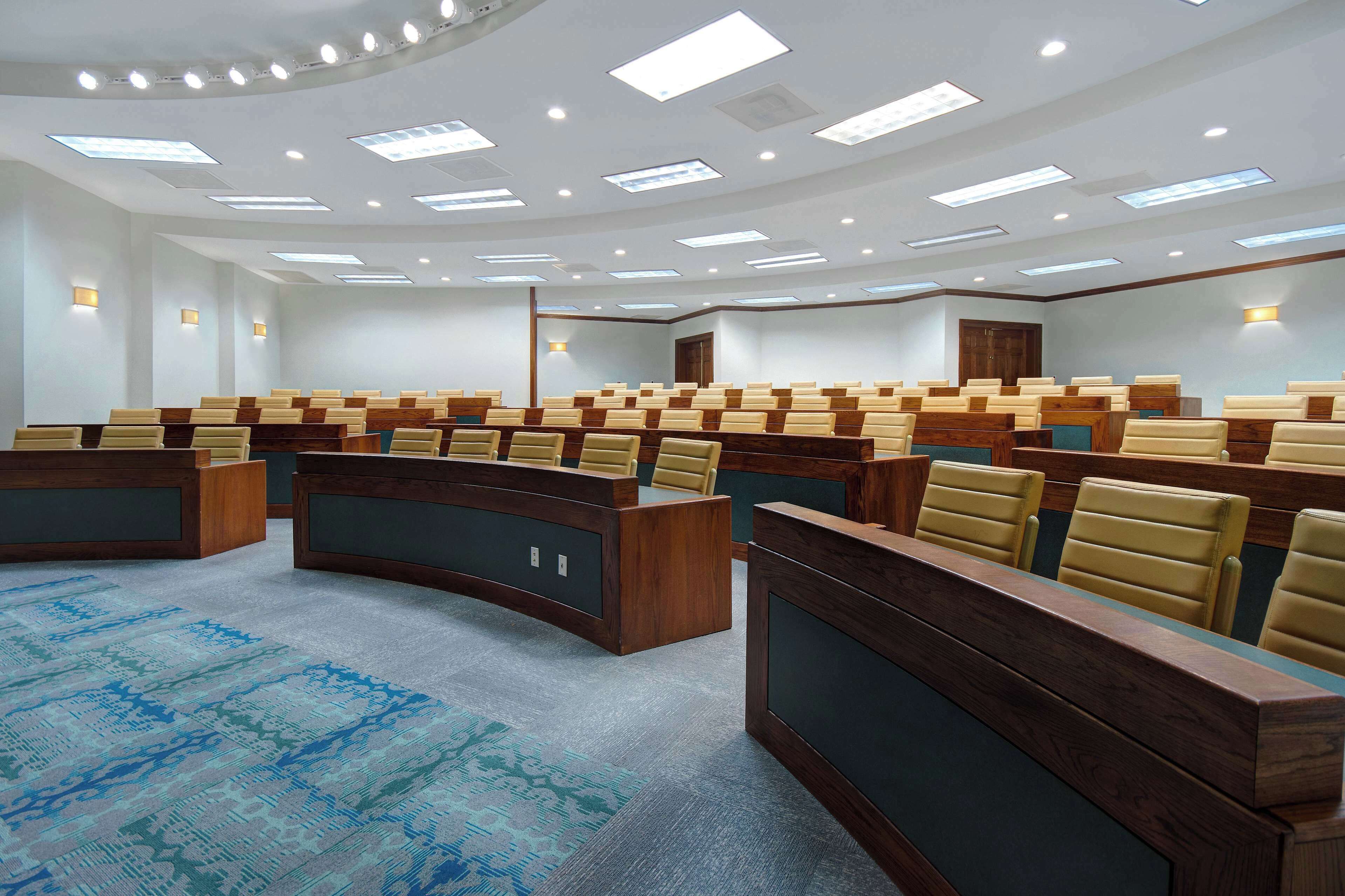 A conference room with rows of wooden desks and beige chairs, equipped with overhead lighting and a patterned carpet.